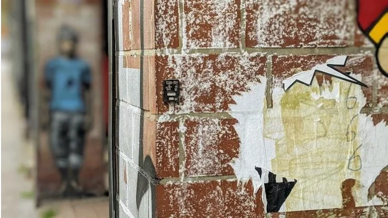 Eine Backsteinwand, voller weißer farbe und einer kleinen, schwarzen Box. Auf dieser Box befindet sich ein Graffito.