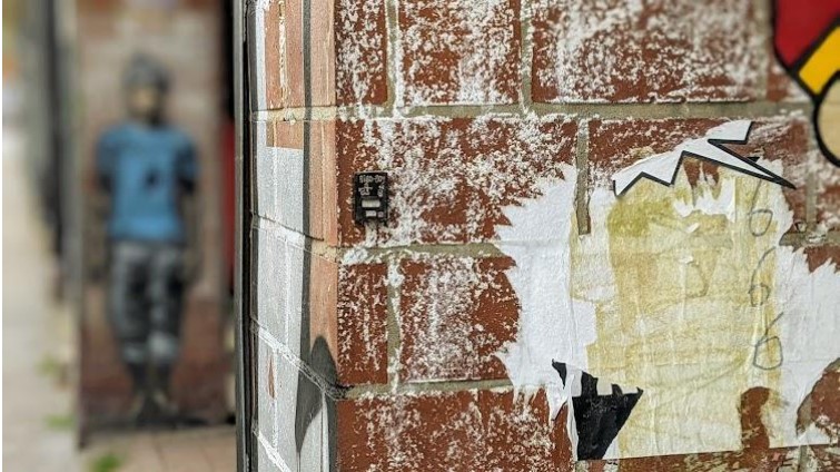 Eine Backsteinwand, voller weißer farbe und einer kleinen, schwarzen Box. Auf dieser Box befindet sich ein Graffito.